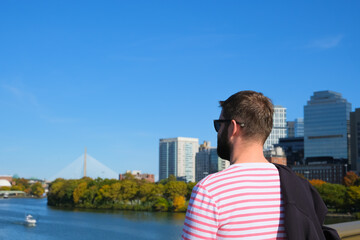 man stands by the river, gazing at the Boston skyline on a clear autumn day. The iconic bridge and vibrant cityscape are visible in the background, reflecting a peaceful and scenic moment.