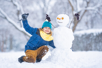 Fun Kid in winter park on a snowy cold winter day. Cute child playing with snow in winter. Portrait of funny child play with snowman. Winter activities Christmas. Kid building snowman in snowy park.