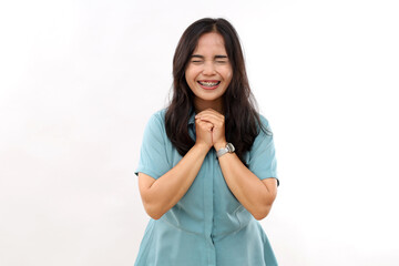 Grateful positive Asian woman clasps hands has beauty face smiles toothily keeps eyes closed dressed in casual casual shirt anticipates something very pleasant isolated over white background