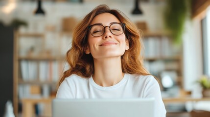 A smiling woman in glasses enjoys a moment of relaxation at her desk with a laptop, conveying a sense of satisfaction and contentment in a bright office.