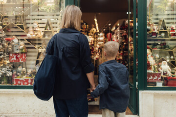 Fototapeta premium Cute little school kid boy with mother on Christmas market. Mom and son look in the window of a gift shop and choose surprises for the family. Holidays, Christmas, childhood and people concept