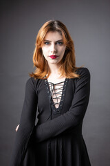Fototapeta premium Red-haired gothic girl in a black dress on a gloomy background in the studio.