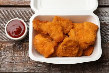 Delicious chicken nuggets in takeaway container and ketchup on wooden table, above view