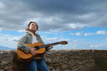 Young woman playing guitar outdoors under cloudy sky