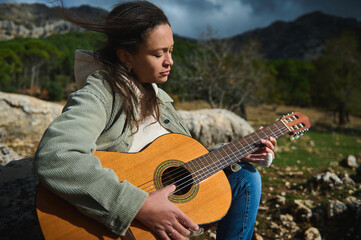 Woman playing guitar outdoors in mountain landscape with green jacket