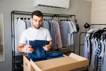 Latin man putting clothes in a cardboard box while organizing his walk-in closet full of shirts and...
