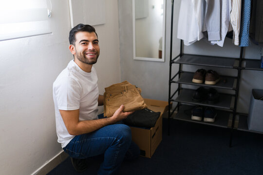 Hispanic man happily arranges clothes in a cardboard box while holding a pair of pants