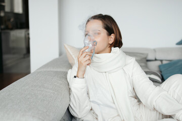 Young Woman Using Nebulizer Mask For Inhalation, Sitting On Sofa At Home, Treating Respiratory Illness