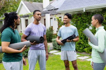 Young multiracial friends holding yoga mats, chatting and smiling outdoors in garden