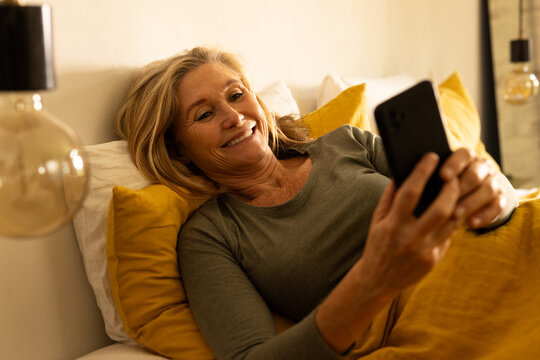 senior woman relaxing in bed, smiling while using smartphone at home