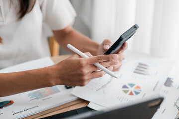 Close-up of hands using a smartphone and stylus to review financial charts and graphs on paper in a bright office.