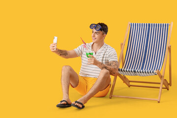 Young man with cocktail and deck chair taking selfie on yellow background