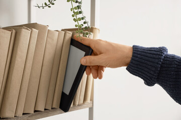 Young man taking e-reader from shelf with books at home, closeup
