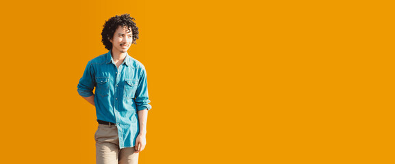 A young man with curly black hair wearing a casual denim shirt and light beige pants poses against...