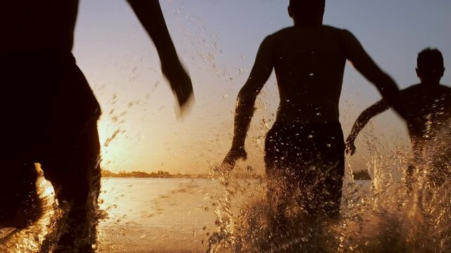 Silhouettes of group of four happy young friends running into lake having fun splashing in water enjoying freedom, adventure on summer holiday at sunset