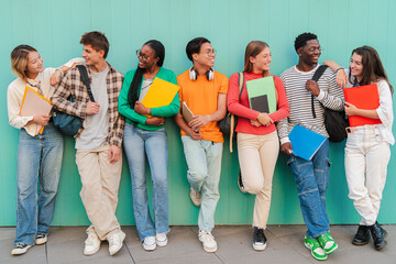 Diverse Group of College or High School Students Share Cheerful Moments Between Classes, Carrying Colorful Folders and Backpacks While Leaning Against Vibrant Teal Wall, Teenage Classmates talking