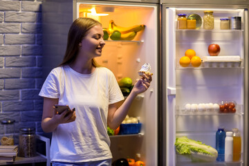 Young woman with mobile phone and cake near open fridge in kitchen at night