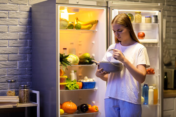 Young woman with spoiled food near open fridge in kitchen at night