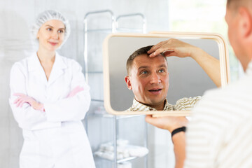 Obraz premium Satisfied middle-aged man patient holding big mirror sitting on clinical chair in aesthetic clinic