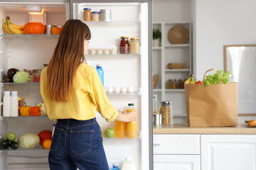 Young woman taking juice from open fridge in kitchen, back view