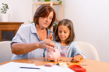 Senior woman and young girl insert coins into piggy bank on table, creating heartwarming moment of connection and shared financial wisdom at home 