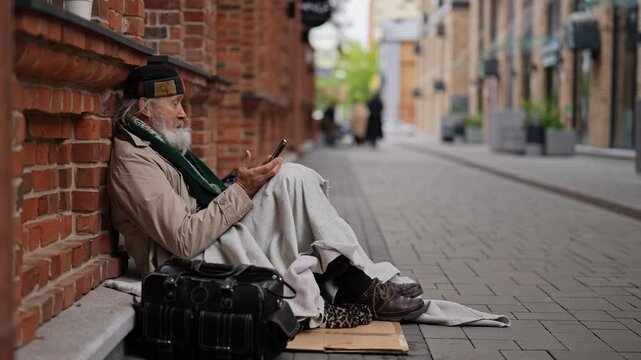 Frozen elderly beggar man using smartphone sitting on cold winter street, wrapped in blanket for warmth, surrounded by meager belongings. Older tramp seek work and government assistance.