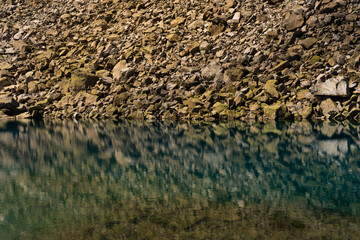 Close-up view of a rocky shoreline perfectly reflected in the calm, clear waters of alpine lake. The image captures the intricate textures of the rocks and the serene mirror-like quality of the water