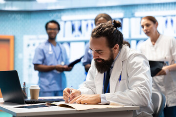 General practitioner writing a prescription for treatment plan at his desk, recommending medicine for disease prevention. Doctor consulting analytics and hospital records in a medical facility.