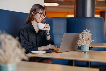 A young woman leisurely sips her coffee while immersed in work on her laptop in a warm, cozy cafe...