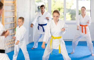 Girl with family, dressed in kimonos, stand in row during class repeat hand movements and punches after man teacher. Experienced male instructor conducts personal karate training for large family