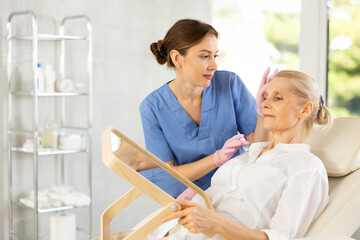 Obraz premium Aged woman having consultation about face aesthetic with a specialist while looking in the mirror on clinical chair