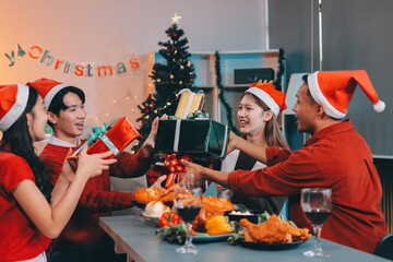 Group of young Asian man and women as friends having fun at a New Year's celebration, holding gift boxes standing by Christmas tree decoration, midnight countdown Party at home with holiday season.
