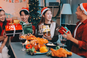 Group of young Asian man and women as friends having fun at a New Year's celebration, holding gift boxes standing by Christmas tree decoration, midnight countdown Party at home with holiday season.