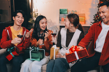 Group of young Asian man and women as friends having fun at a New Year's celebration, holding gift boxes standing by Christmas tree decoration, midnight countdown Party at home with holiday season.