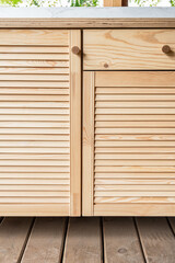 Wooden cabinets of outdoor kitchen with louvered doors, drawers, wooden handles and plywood countertop, stand on a wooden terrace close-up view