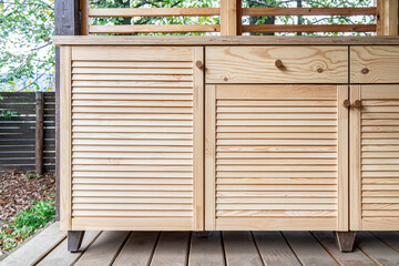 Wooden cabinets of outdoor kitchen with louvered doors, drawers, wooden handles and plywood countertop, stand on a wooden terrace