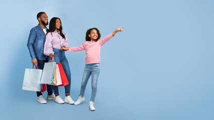 Family Shopping Concept. Full length body size portrait of African American cheerful people holding shopping bags with new clothes, excited little girl pointing aside at copy space, blue studio wall