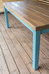 Dining table is made of pine wood with blue legs close-up view. Massive table on terrace with wooden flooring and has a view of the nearby grassy area