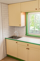 Fragment of kitchen with yellow cabinets with wooden handles features a plastic countertop with a marble-like finish, and upper cabinets installed around the window