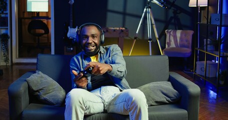 Enthusiastic gamer on couch using controller and headphones to play videogame in neon lit apartment. Player in RGB illuminated living room using high tech gaming console joystick, camera B