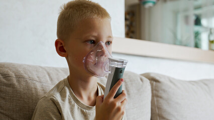 A young boy sits quietly at home, using an inhaler for treatment. This is a common method for managing asthma and preventing respiratory illnesses like colds.