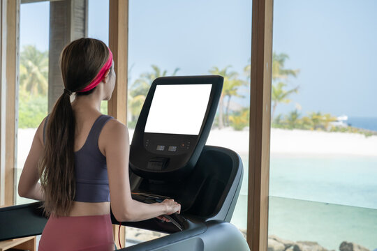 Woman is working out in gym. Doing cardio training on treadmill with white screen mockup , large windows with ocean view raining outside.