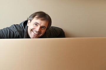 A cheerful man peeks over a large cardboard box, showcasing excitement and joy in a simple yet engaging moment.