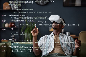 Young African American man wearing VR headset sitting on sofa in living room coding in virtual app