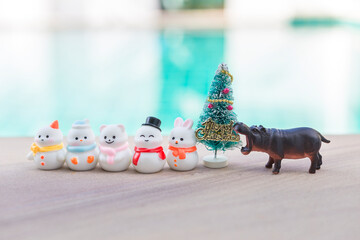 Cute little snow man collections with Hippopotamus toy and Christmas tree over blurred swimming pool background, outdoor day light, Christmas concept background idea