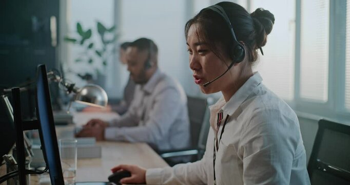 Call center office: Portrait of friendly Asian female hotline operator talking on headset, providing online customer assistance, looking at camera. Multiethnic team of technical support specialists.