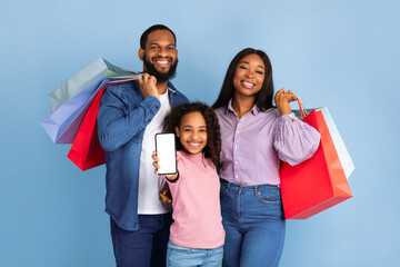 Obraz premium Portrait of happy family of three people of color holding shopping bags, cheerful girl showing smartphone with white empty screen in hand, isolated on blue studio background, mock up, blank space