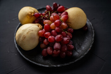 Clusters of red grapes and yellow pears on a black mica plate on a dark background.