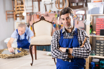 Obraz premium Middle-aged male professional craftsman, dressed in overalls, poses in working atmosphere of restoration workshop after working with antique furniture