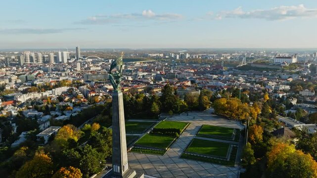 Aerial view of the Slavin Memorial downtown and castle in Bratislava with the cityscape stretching beneath it, framed by vibrant autumn foliage and clear skies.
